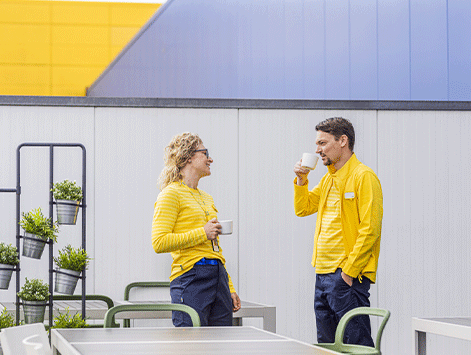Dos empleados de tienda con uniformes amarillos y azules parados en una terraza exterior, charlando y sonriendo mientras toman café en tazas blancas.