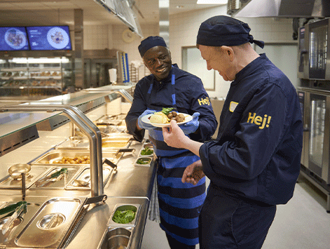 Dos sonrientes empleados de cocina con uniformes azul oscuro que llevan la palabra "Hej!" en la manga comparten un plato de comida junto al mostrador de una cafetería.