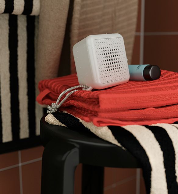 A VAPPEBY portable bluetooth speaker in white placed on folded rust-coloured towels on a black stool in a bathroom.