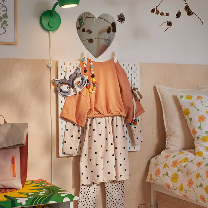 A beige and orange child’s outfit hangs on a SKÅDIS pegboard topped by a HOLMÖARNA heart-shaped mirror.