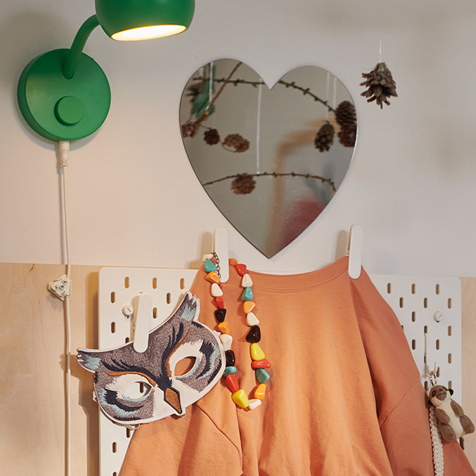 A beige and orange child’s outfit hangs on a SKÅDIS pegboard topped by a HOLMÖARNA heart-shaped mirror.