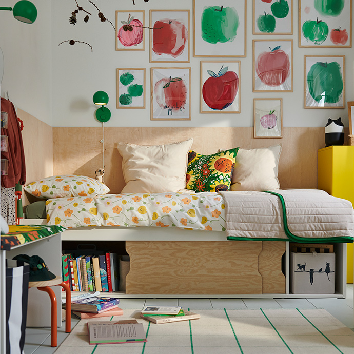 Un dormitorio con una estructura de cama KLIPPUGGLA blanca, con almacenamiento debajo, decorada con ropa de cama y cojines coloridos con estampados florales.