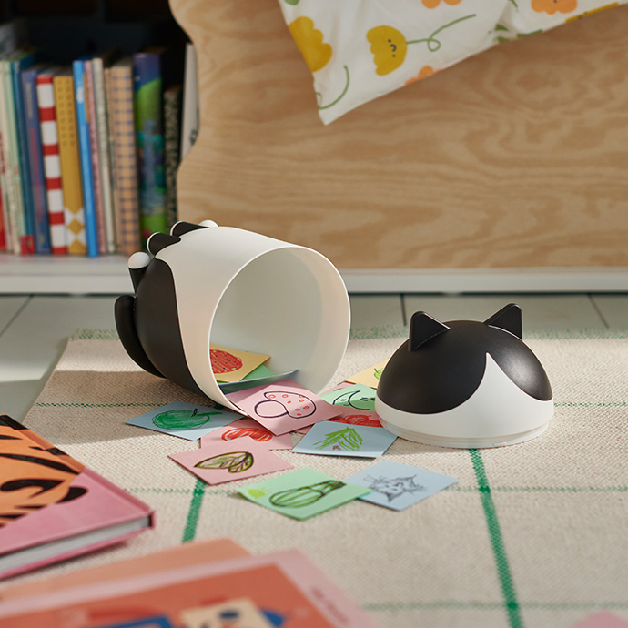 Un contenedor GREJSIMOJS con forma de gato está abierto sobre una alfombra, mostrando su contenido: unas tarjetas de papel de un niño.