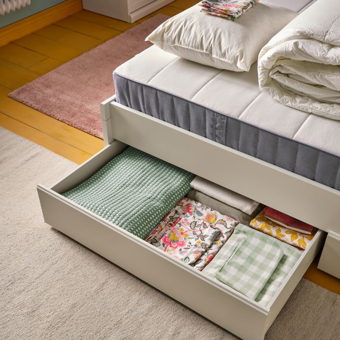 Un dormitorio con una estructura de cama FÅGELFJÄLLET en color blanco roto y cuatro cajas de almacenamiento, una de ellas llena de ropa de cama doblada.