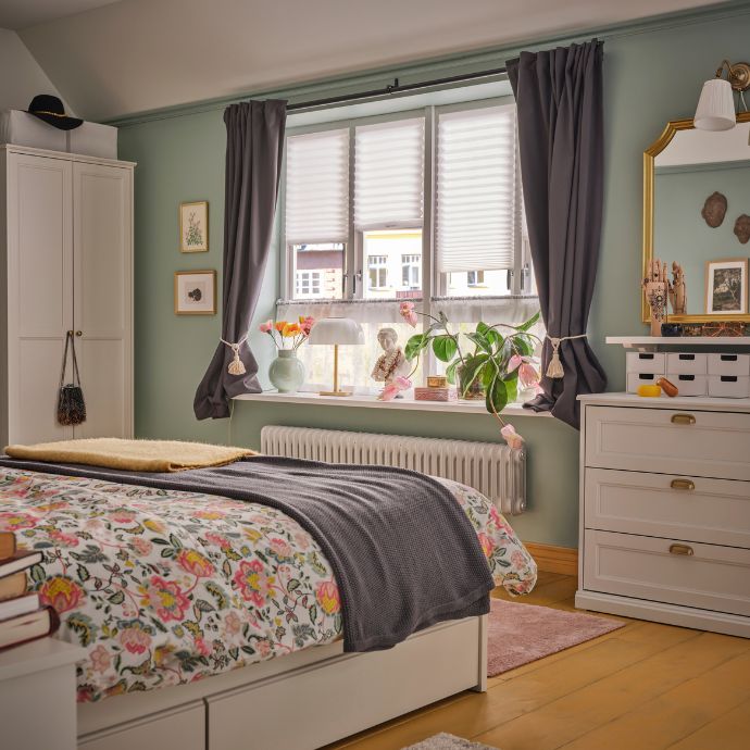 Un dormitorio con cama de sábanas florales y un taburete BEKVÄM junto a la ventana. Las cortinas opacas KORGMOTT enmarcan la ventana.