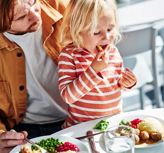 Un niño rubio con camiseta de rayas come una patata en una mesa soleada junto a su padre, ante un plato de albóndigas suecas y puré de patatas.