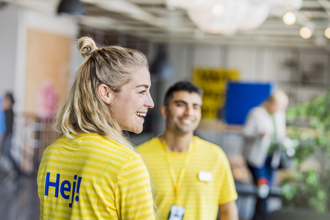 Primer plano de una colaboradora de IKEA sonriente con el pelo recogido, vistiendo una camiseta de rayas amarillas con el texto "Hej!" en azul y un compañero al fondo.