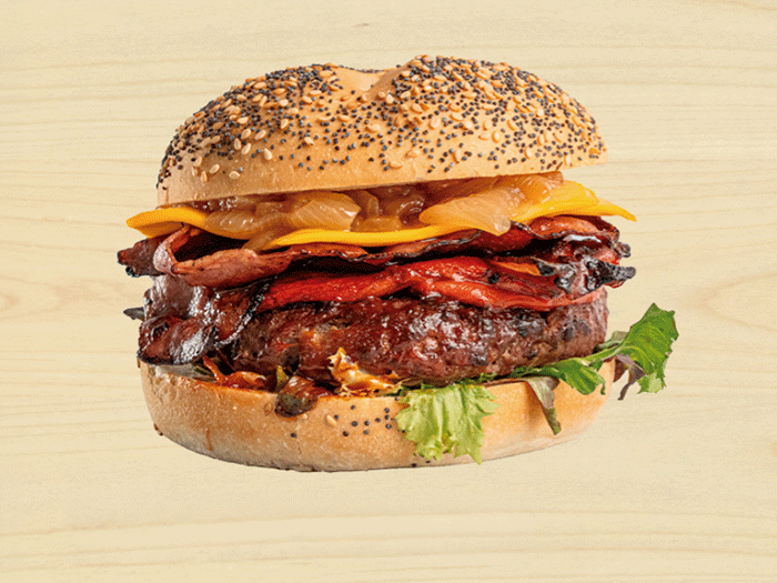 Una hamburguesa gourmet con queso cheddar fundido, bacon crujiente, cebolla caramelizada y lechuga en pan con semillas de amapola sobre fondo de madera clara.