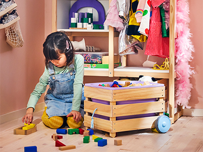 Young girl playing on the floor with wooden blocks next to a toy box with wheels.