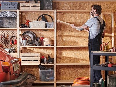 Man organizing wooden boards on workshop shelving near tools and a classic car.