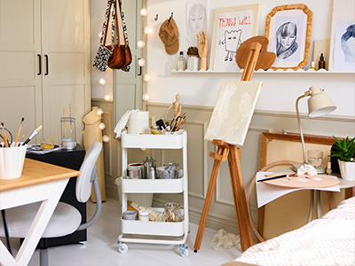 Art studio setup with a wooden easel, white utility cart with supplies, and desks.