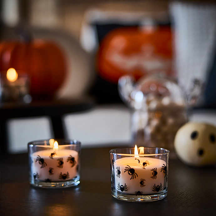 Two lit scented KUSTFYR candles with spider designs on glass placed side by side on a table.