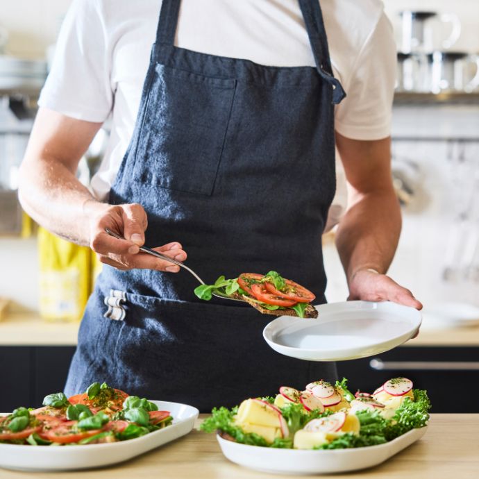 Barista plates open sandwiches topped with greens and tomato onto dish; salads and prepared meals arranged on counter.