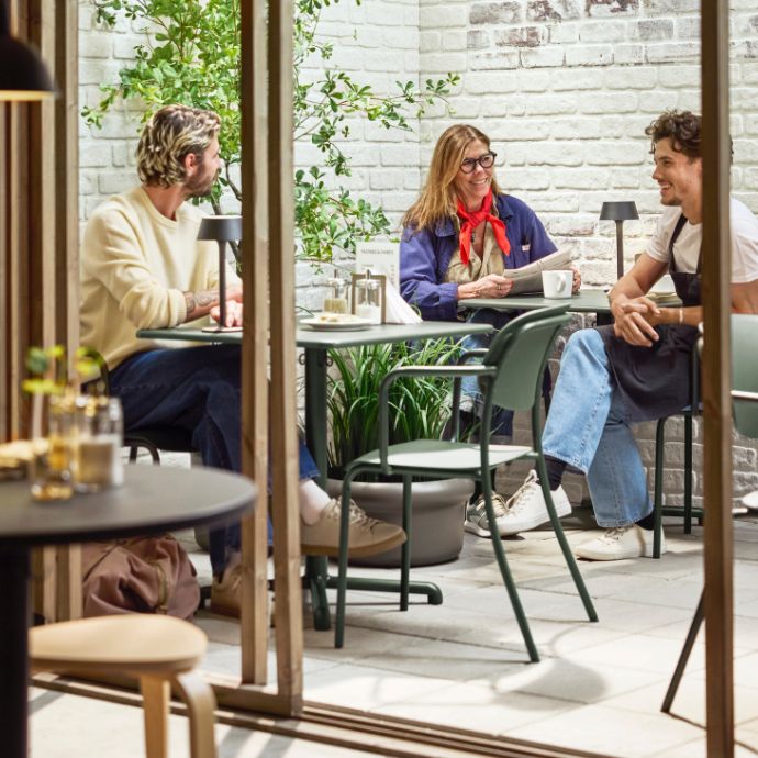 Three people sit outdoors at café table with drinks, talking and reading menu; greenery, brick wall, and glass doors visible.