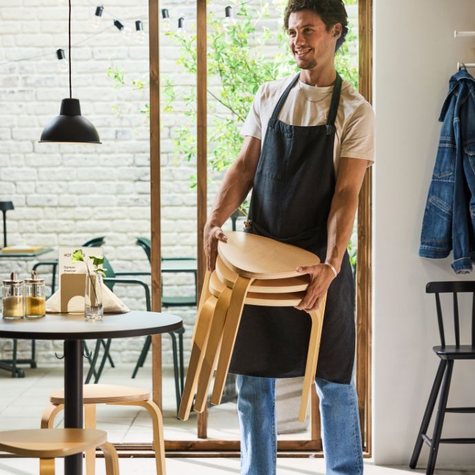 Barista in apron carries stacked stools inside café, arranging furniture near table, window and hanging lamp in bright café.