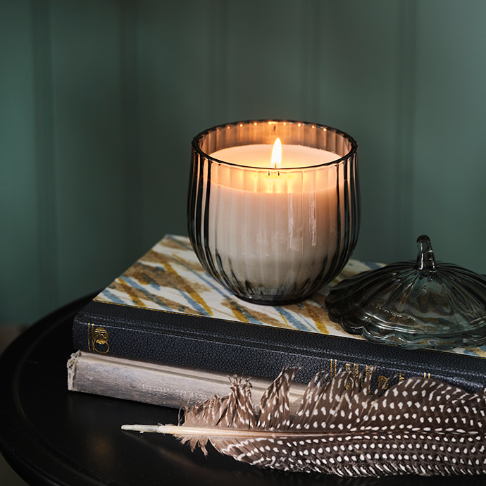 A SNÖSISKA scented candle with a glass lid stands on a stack of books. In front of the books lies a feather.