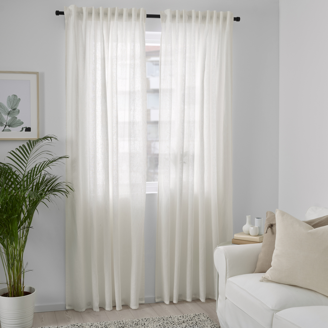 Semi-sheer white curtains hanging on a black rod in front of a window, set in a living room with a white sofa and a houseplant.