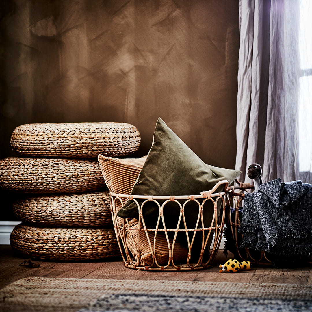 Rustic decorative corner featuring stacked woven floor cushions, a rattan basket filled with throw pillows, and blankets near a window.