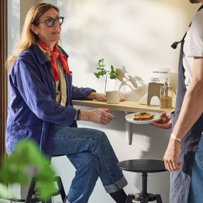 Customer seated at café counter receives plated sandwich from barista; cup, plant, and condiments on table in bright setting.