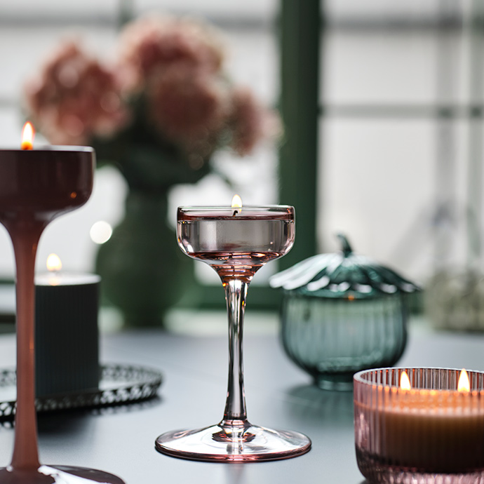 A BLOMSTER candle holder in pale pink glass with a lit candle in it stands on a dark table surrounded by other candles.