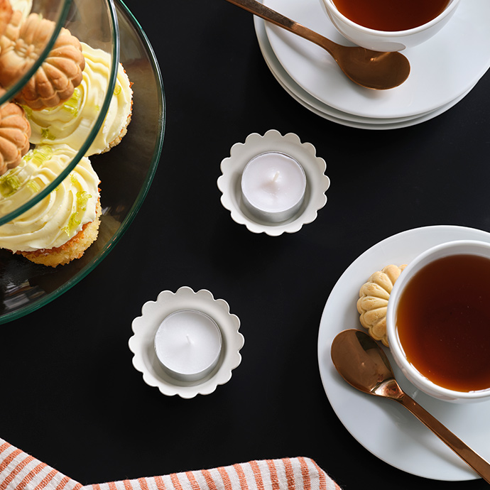 Two SNÖSISKA candle holders in off-white stand on a black table surrounded by two cups of tea and a cake stand with cookies.