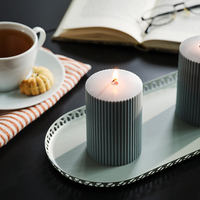 A dark grey-green SNÖSISKA pillar candle stands on candle dish on a black table with a book and a cup of tea.