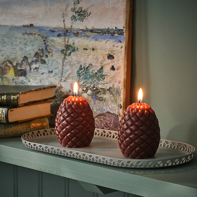 A couple of SNÖSISKA pillar candles shaped like pine cones stand on a pale grey-green SNÖSISKA candle dish on a green shelf.
