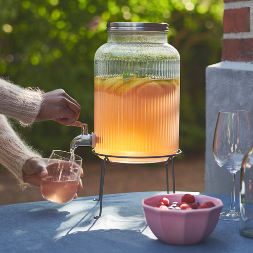 La persona vierte la bebida de naranja del dispensador VARDAGEN en un vaso. En el exterior, la mesa tiene fresas y dos vasos.