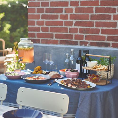 Mesa al aire libre con mantel azul, varios platos, bebidas y un soporte de jarra varden que sostiene un recipiente de vidrio con un grifo, colocado contra una pared de ladrillo rojo.