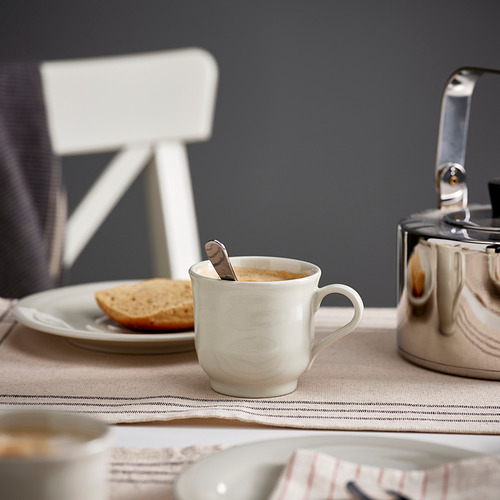 Una taza de cerámica blanca llena de café descansa sobre un mantel beige cerca de una tetera metálica y un pequeño plato con tostadas.