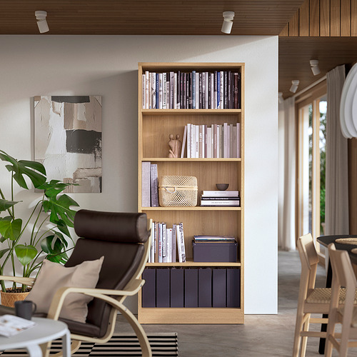 Estantería de roble con libros y decoración, junto a silla oscura, planta y pintura moderna.