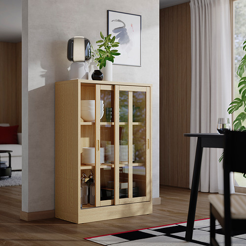 Mueble de roble con frente de vidrio con platos, botellas, plantas, al lado de la silla, cerca de la ventana.