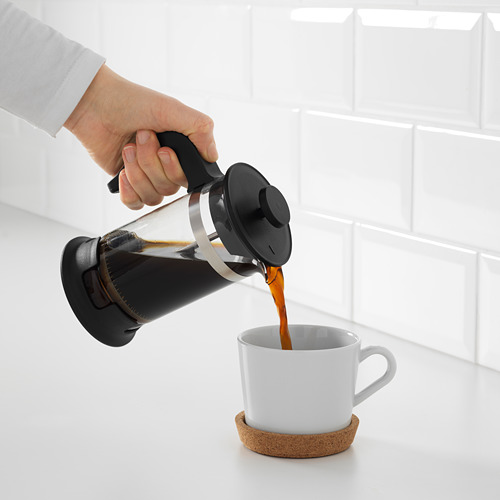 Hand vierte café de una prensa francesa negra en una taza blanca sobre un posavasos de corcho contra una pared de azulejos blancos.