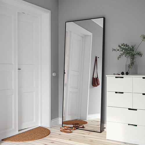 El espejo Hövet se apoya en la pared, reflejando el pasillo. El marco negro complementa la decoración moderna, rodeado de suelo de madera y bolso rojo.