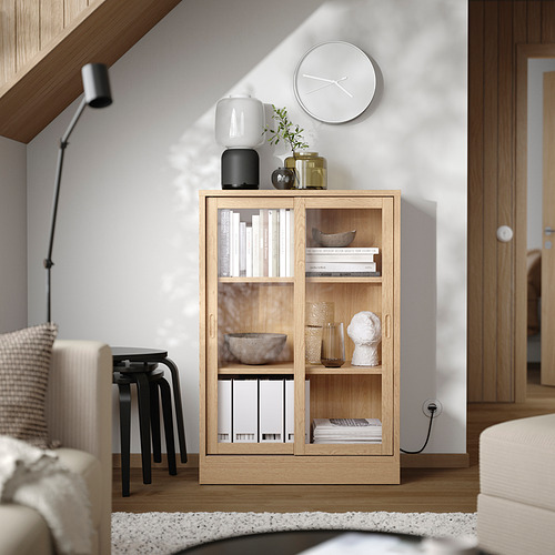 Mueble TONSTAD de madera en roble claro con puertas correderas de cristal, estanterías con libros, cuencos y jarrones, reloj blanco y decoración en la parte superior.