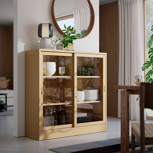 Mueble TONSTAD de roble claro, puertas de cristal, tres estantes. Guarda platos, botellas. Muestra un diseño clásico. Alto, encaja bien en la habitación con otros muebles.