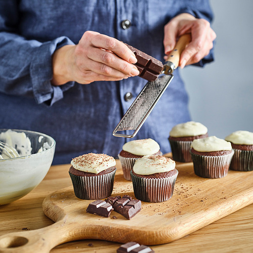 La persona ralla chocolate sobre cupcakes glaseados en una tabla de madera para decorar. Un cuenco de glaseado está cerca.