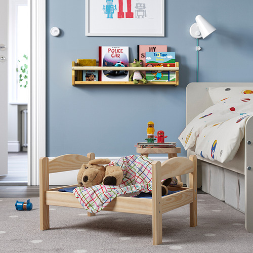 Cama infantil de madera DUKTIG, azul, con oso y manta. Cerca de un estante con libros, una mesita de noche pequeña y una cama blanca más grande. Acogedor, pequeño y colorido.