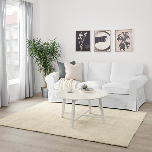 Una sala de estar minimalista con una alfombra ENGELSBORG beige, un sofá blanco, una mesa de centro y una planta verde.