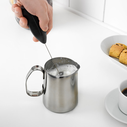 La mano sostiene un espumador negro que hace espuma en una jarra de metal sobre un mostrador blanco, junto al café y las galletas.