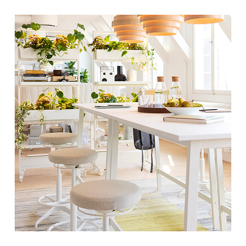 Espacio de trabajo luminoso y bien iluminado con mesas, taburetes y estanterías blancas. Vegetación y artículos de decoración expuestos. Luz natural por las ventanas.