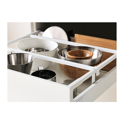 Un cajón de cocina blanco abierto, que contiene cuencos metálicos, un cuenco de madera y una tabla de cortar, sostenidos por compartimentos ajustables.