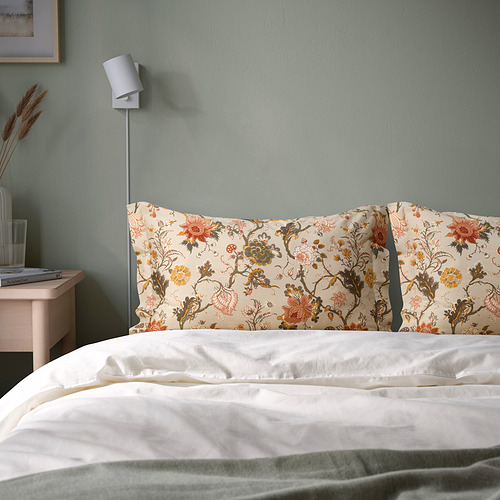 Cama con almohadas florales, lámpara, jarrón y libros. Almohadas NÄSSELKLOCKA en beige contra una pared verde.
