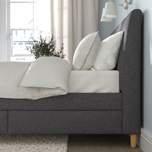 Cama en tonos neutros con cabecero tapizado, almohadas blancas y manta doblada. Cajones de cierre suave en la parte inferior para almacenamiento.