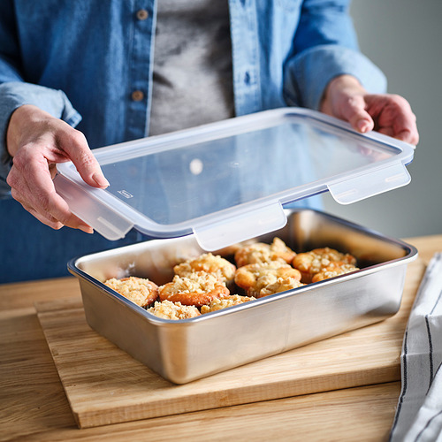 Persona que coloca una tapa transparente a prueba de fugas en un recipiente rectangular de alimentos lleno de productos horneados, lo que garantiza la frescura y evita derrames.