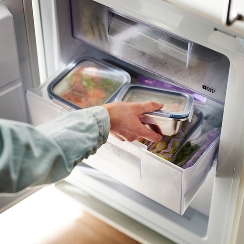 Persona colocando un recipiente de acero inoxidable para alimentos con tapa en un refrigerador. El recipiente es transparente y está diseñado para almacenar las sobras.