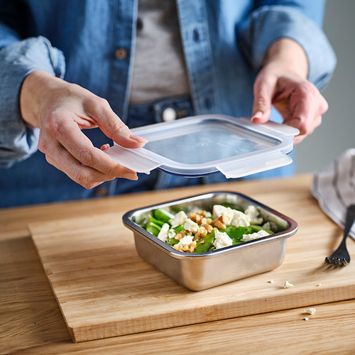 La persona con camisa azul abre la tapa transparente del recipiente metálico para alimentos con ensalada, lo que demuestra un fácil almacenamiento y una función segura de cierre y presión.