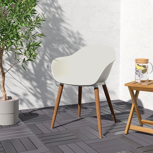 Escena exterior moderna: silla GRÖNSTA blanca con patas de madera junto a una pequeña mesa que sostiene una bebida, cerca de una gran planta contra una pared ligera.