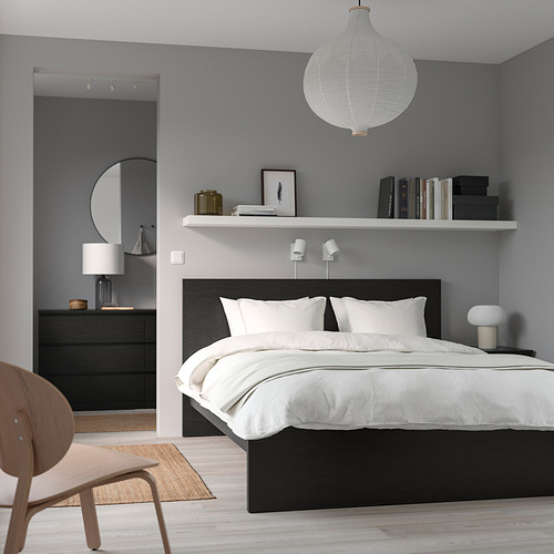 Dormitorio minimalista con paredes grises, cama blanca, tocador oscuro, lámpara redonda, estantería y silla de madera. La cama es baja, más grande que la cómoda.