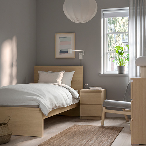 Cama MALM de madera con sábanas blancas, alfombra gris y planta en maceta junto a una ventana luminosa.